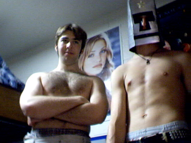 Two shirtless college-age men in a dorm room, one wearing a homemade mask/helmet on his head, the other with arms crossed